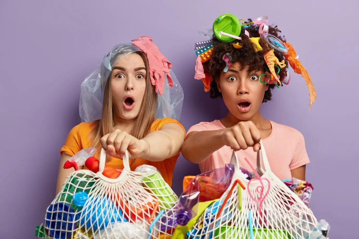 Freigestellte Aufnahme von verblüfften, multiethnischen Frauen, die mit aufgerissenen Augen vor Überraschung starren, Netztaschen mit Plastikmüll halten, wiederverwendbaren Müll recyceln wollen und dicht beieinander vor einer violetten Wand stehen.